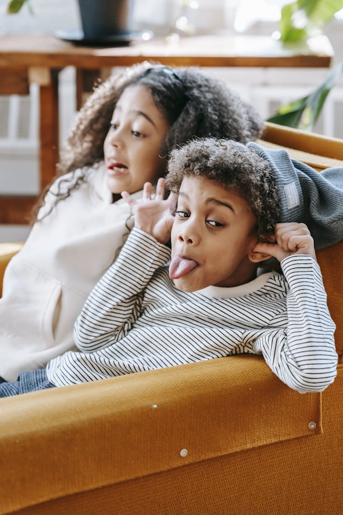 Cheerful little African American children in casual clothes sitting on cozy sofa and showing tongues while having fun together during weekend at home